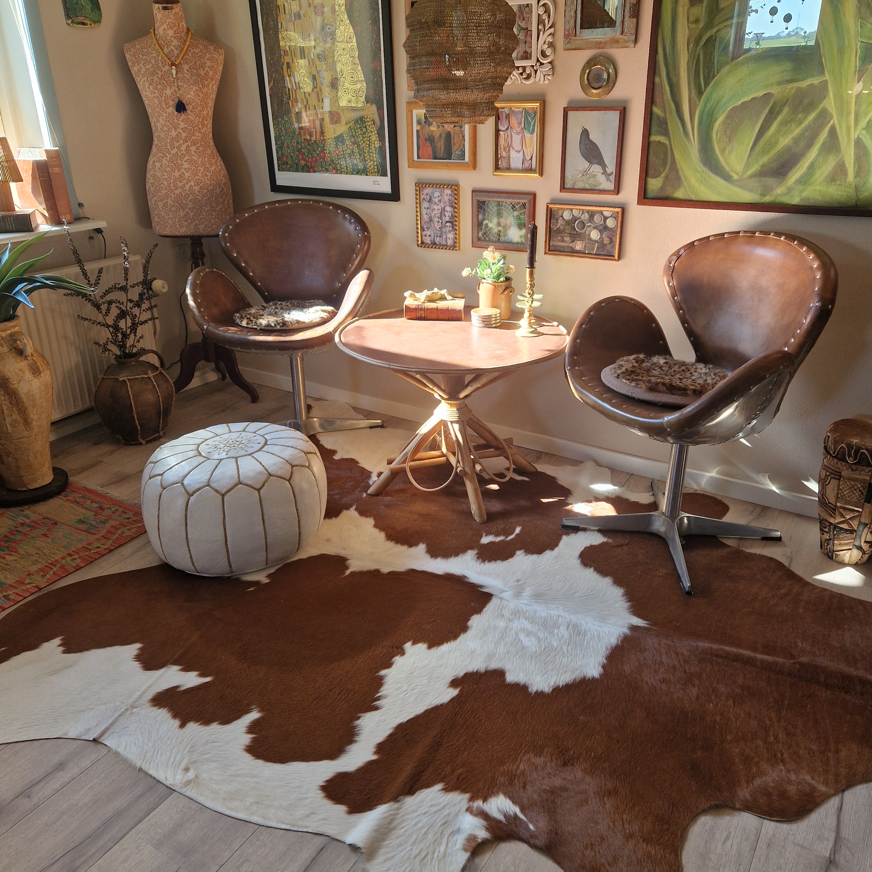 Bruin-witte koeienhuid bij twee leren fauteuils en een ronde tafel in een woonkamer met kunst aan de muur.