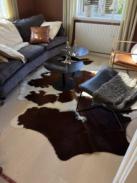 Bruin-witte koeienhuid tussen een donkere bank en fauteuils rond twee zwarte salontafels.