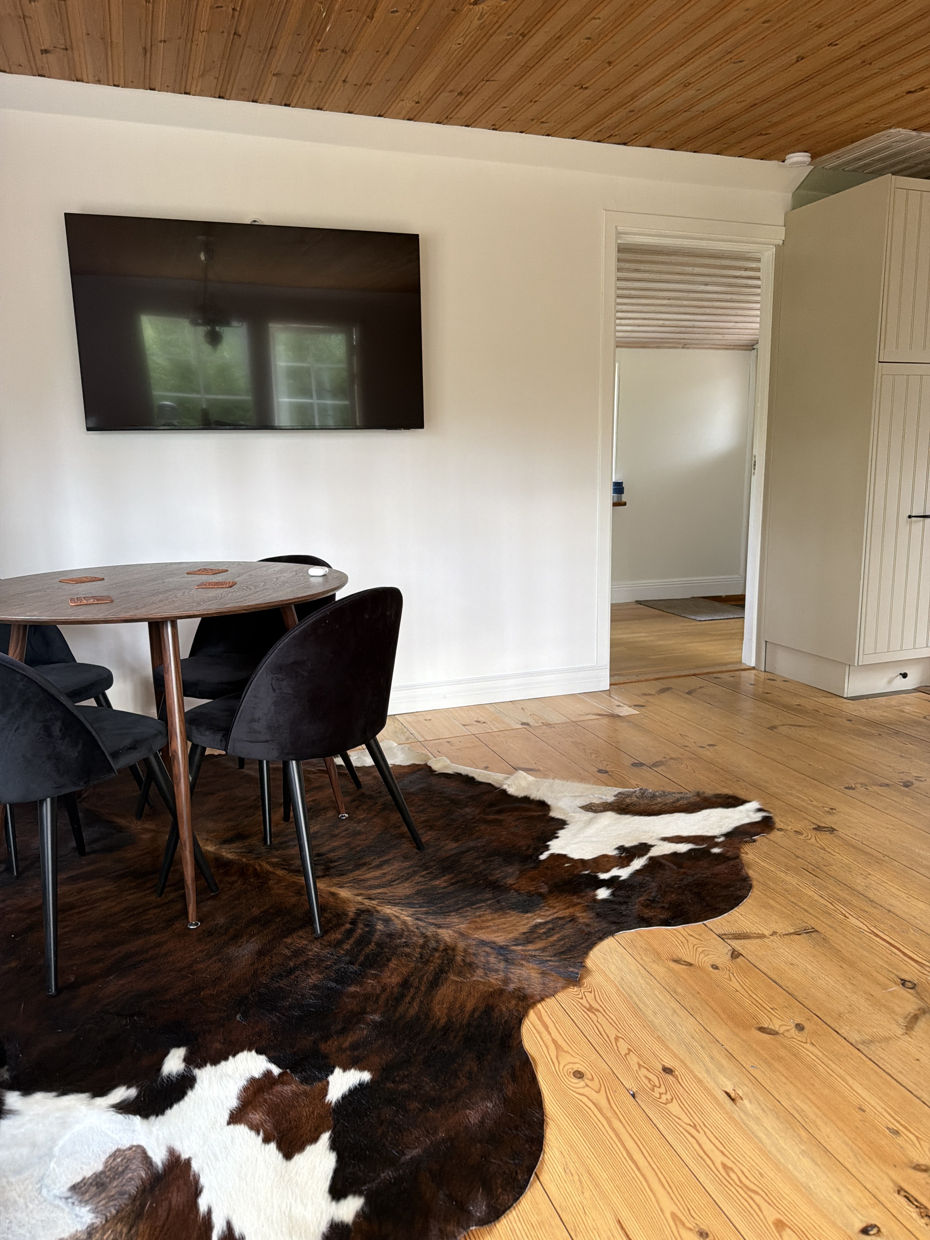 Bruin-witte koeienhuid onder een ronde houten eettafel met zwarte stoelen in een lichte kamer.