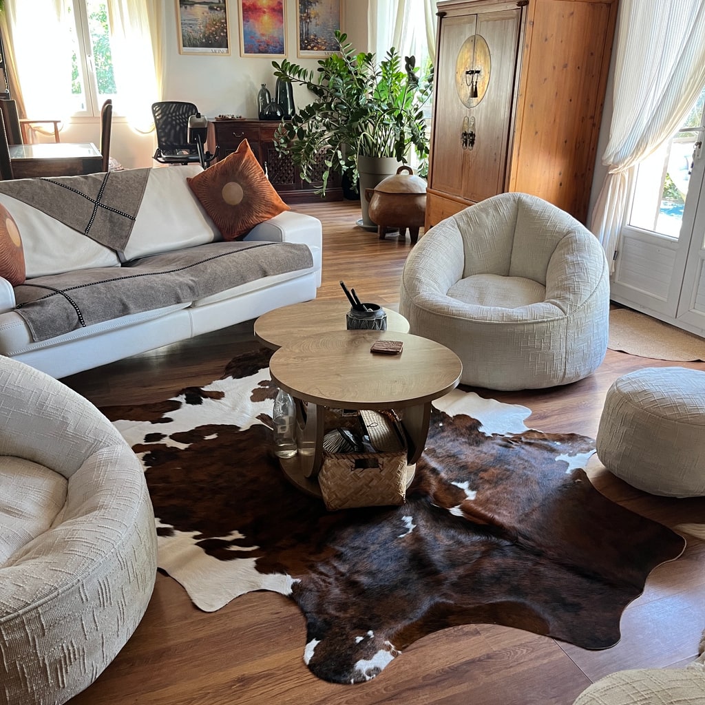 Donkerbruin-witte koeienhuid onder twee houten salontafels in een lichte woonkamer met witte zitmeubels.